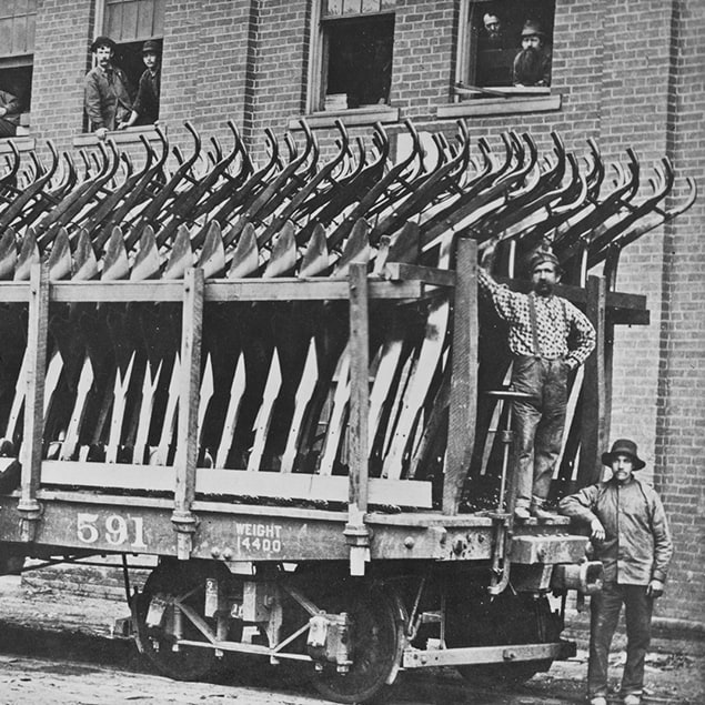 Fotografi fra 1882 av tre menn som står ved siden av en Deere & Co-togvogn lastet med stålploger som er klare til forsendelse, og fabrikkarbeidere som ser ut av vinduet i bygningen bakenfor
