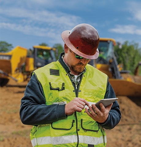 Bygningsarbeider i grønn sikkerhetsvest som ser på mobilen sin, foran en laster og en dumper.