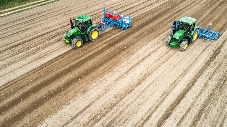 To John Deere&trade; 6M traktorer p&aring; et nakent jordstykke. Den ene drar en Monsomen plantemaskin. Den andre drar en Lemken s&aring;kornbed-kombinasjon