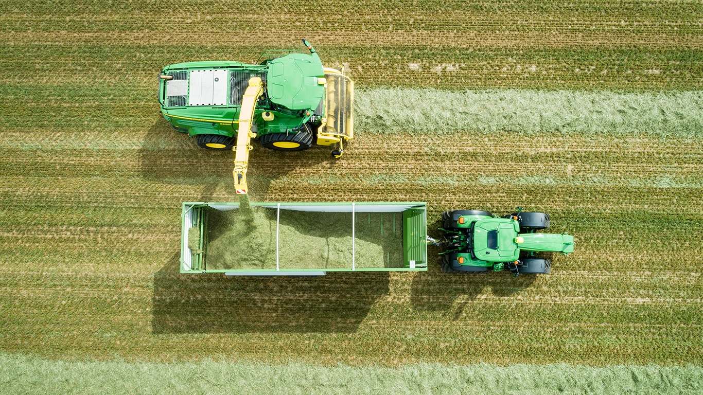 Luftfoto av John Deere&trade; 8000-serie, en selvg&aring;ende forh&oslash;ster med en gress-pickup som losser silof&ocirc;r p&aring; en vogn som trekkes av en John Deere&trade; traktor