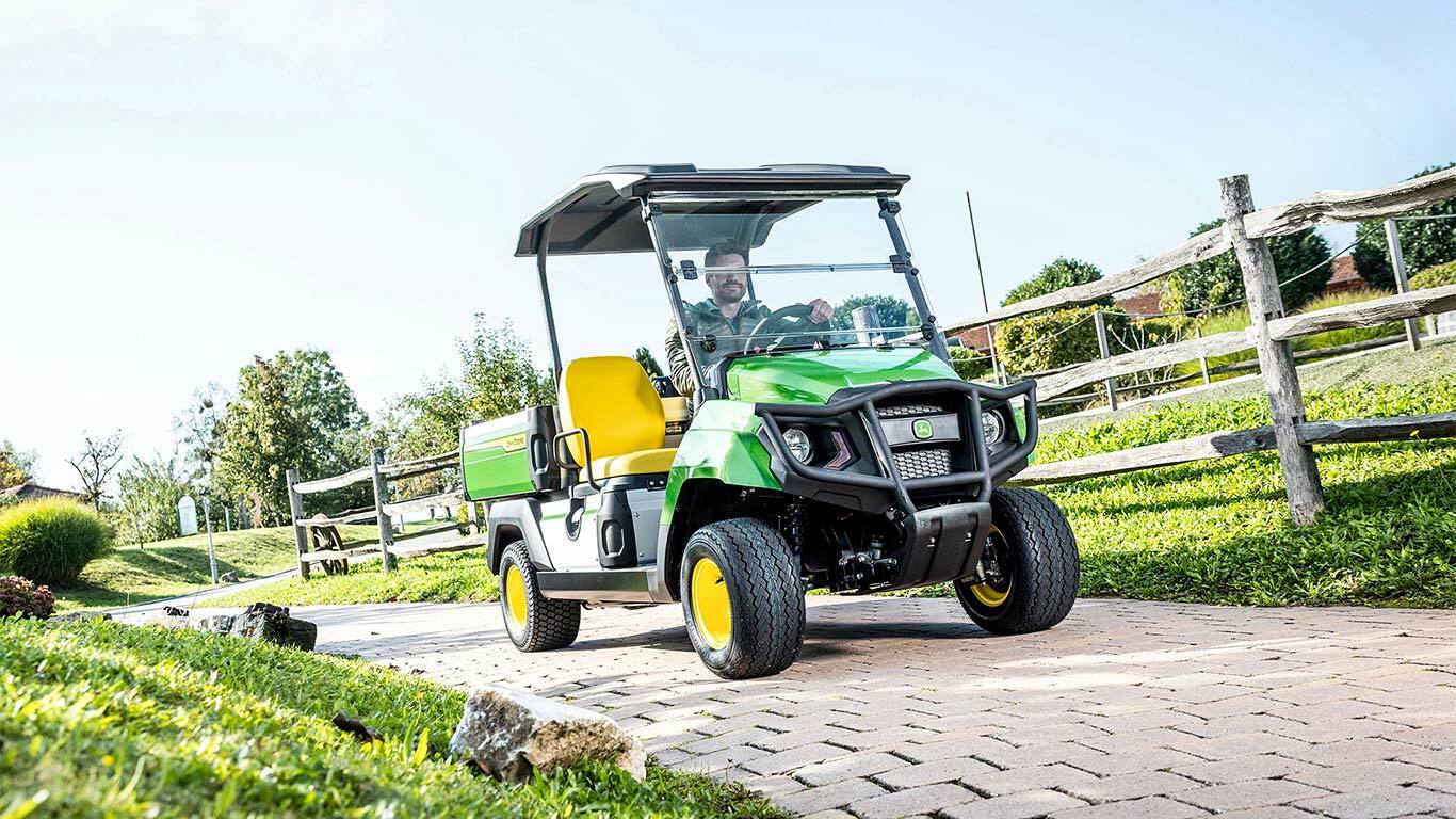 Grønn John Deere Gator GS nyttekjøretøy som kjører på en asfaltert bane gjennom et anlagt område med gress og tregjerder.