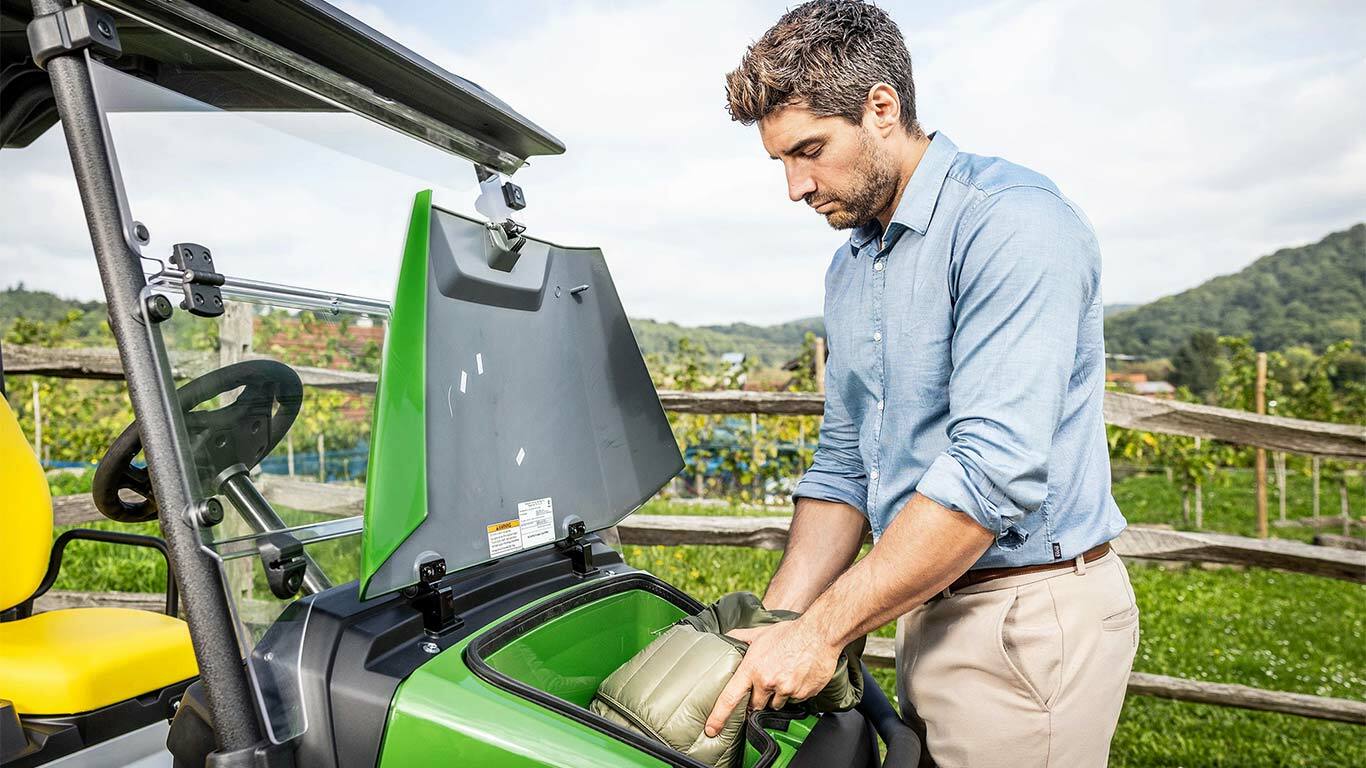 Person som plasserer en polstret pose i oppbevaringsrommet foran på en grønn John Deere Gator GS parkert utendørs nær et tregjerde og en vingård.