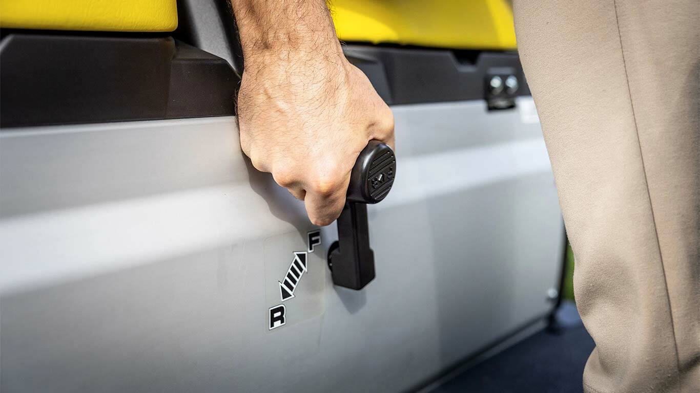 Nærbilde av en hånd som betjener girspaken på en John Deere Gator GS, som viser forover- og reversposisjoner ved siden av gule seter.