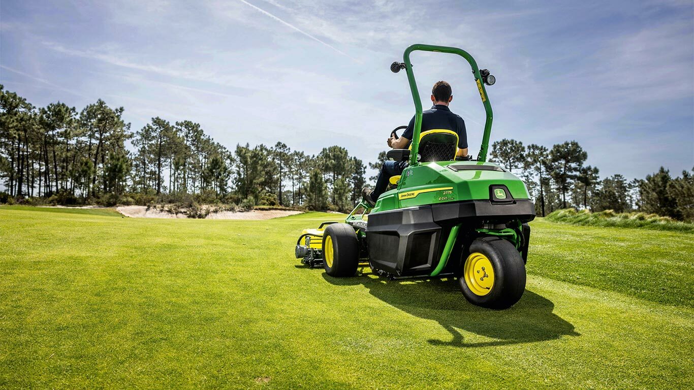 En person kjører en grønn John Deere 2775 E-Cut™ elektrisk gressklipper for green på en godt vedlikeholdt golfbane, med trær og en delvis overskyet himmel i bakgrunnen.