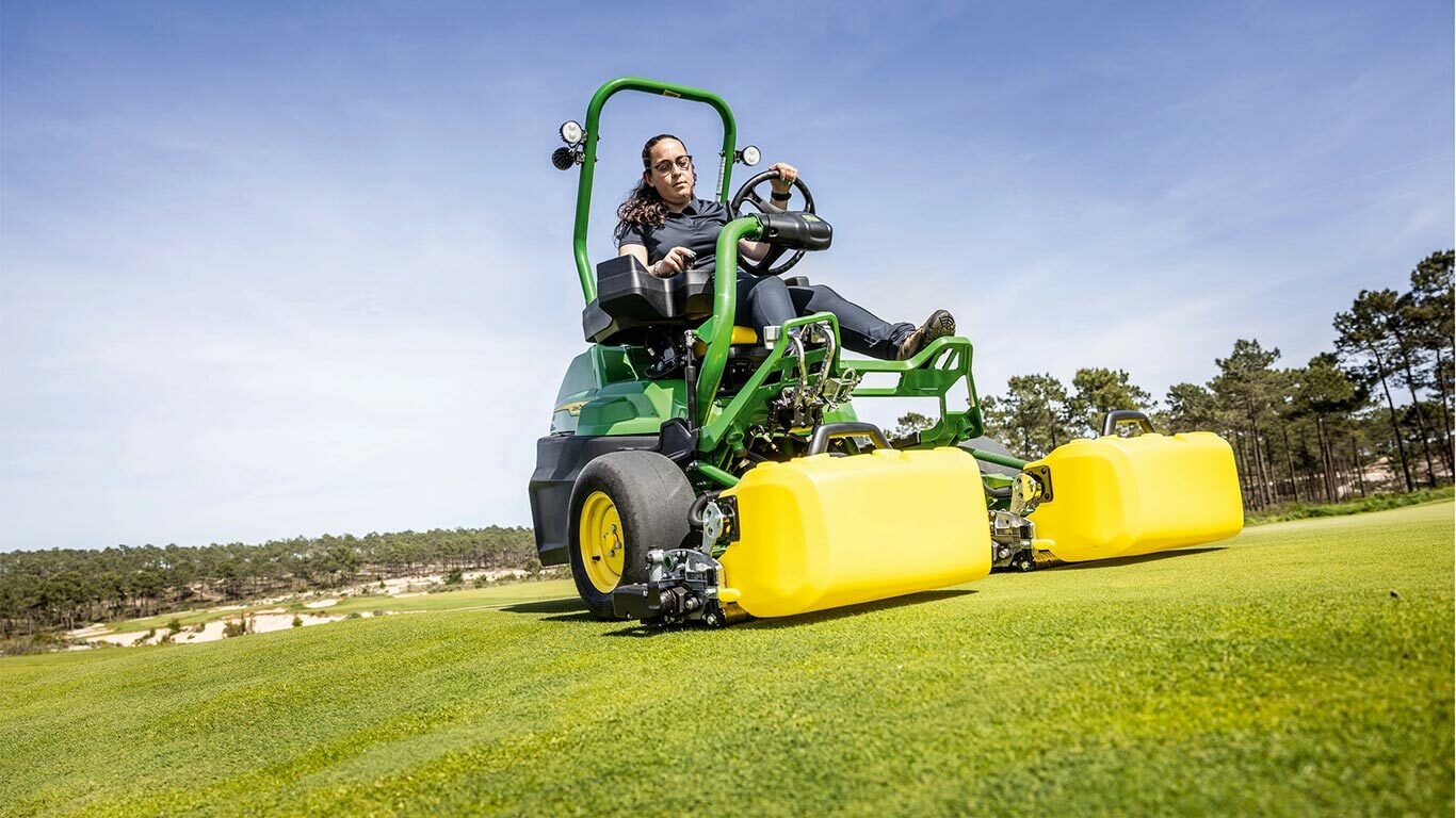 En person betjener en grønn og gul John Deere 2775 E-Cut™ elektrisk gressklipper med frontruller dekket av et gult hus på en golfgreen, med trær og en klar, blå himmel i bakgrunnen.