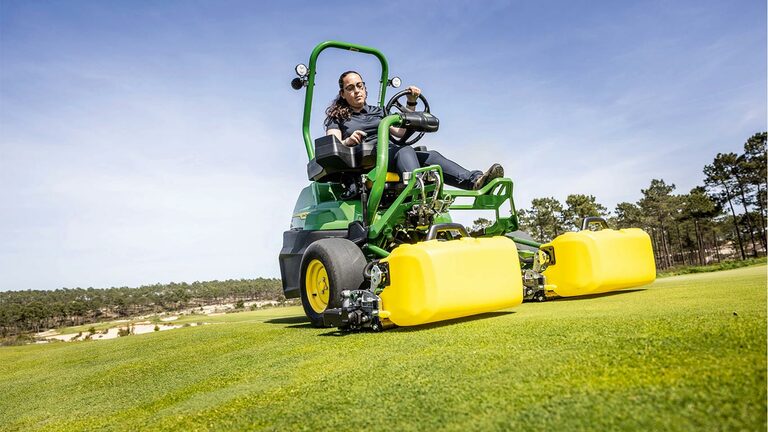 En person betjener en grønn og gul John Deere 2775 E-Cut™ elektrisk gressklipper med frontruller dekket av et gult hus på en golfgreen, med trær og en klar, blå himmel i bakgrunnen.