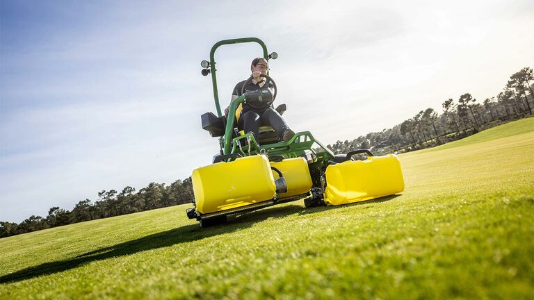 En person betjener en grønn og gul John Deere 2775 E-Cut™ elektrisk gressklipper med tre store frontruller dekket av gule hus på en golfgreen, med trær og en delvis overskyet himmel i bakgrunnen.