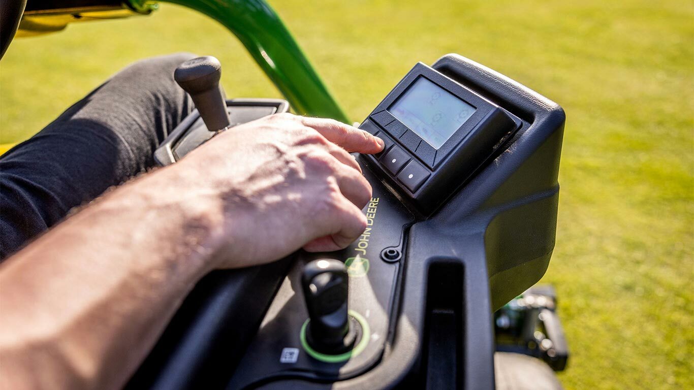 Nærbilde av en persons hånd som betjenter TechControl-panelet på en John Deere 2775 E-Cut™ elektrisk gressklipper, ved å trykke på knappene på et digitalt display med en joystick og andre kontroller synlige.