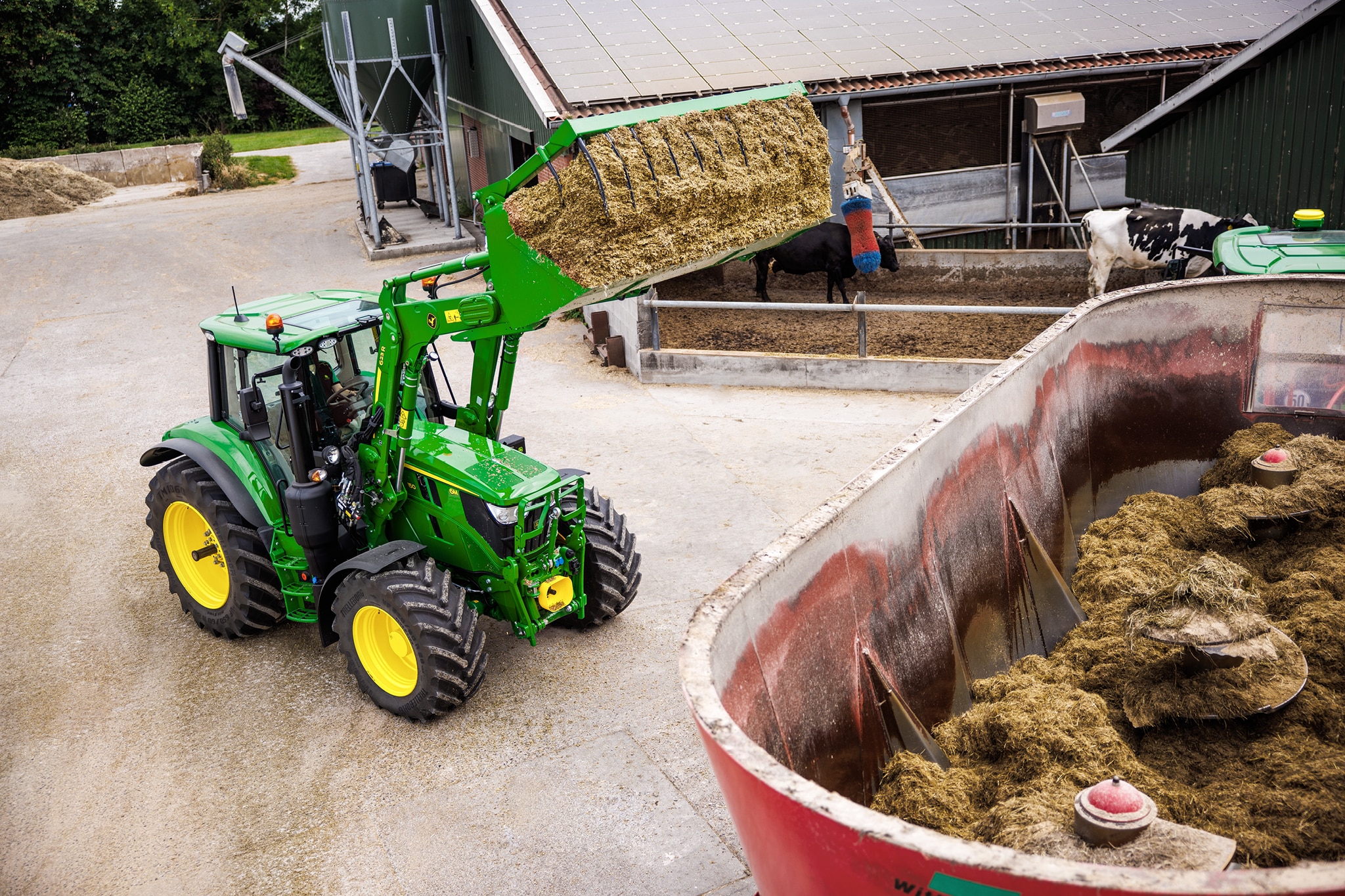 John Deere-traktor i 6M-serien som bruker en frontlaster til å mate silofôr inn i en blandervogn på en gårdsplass.
