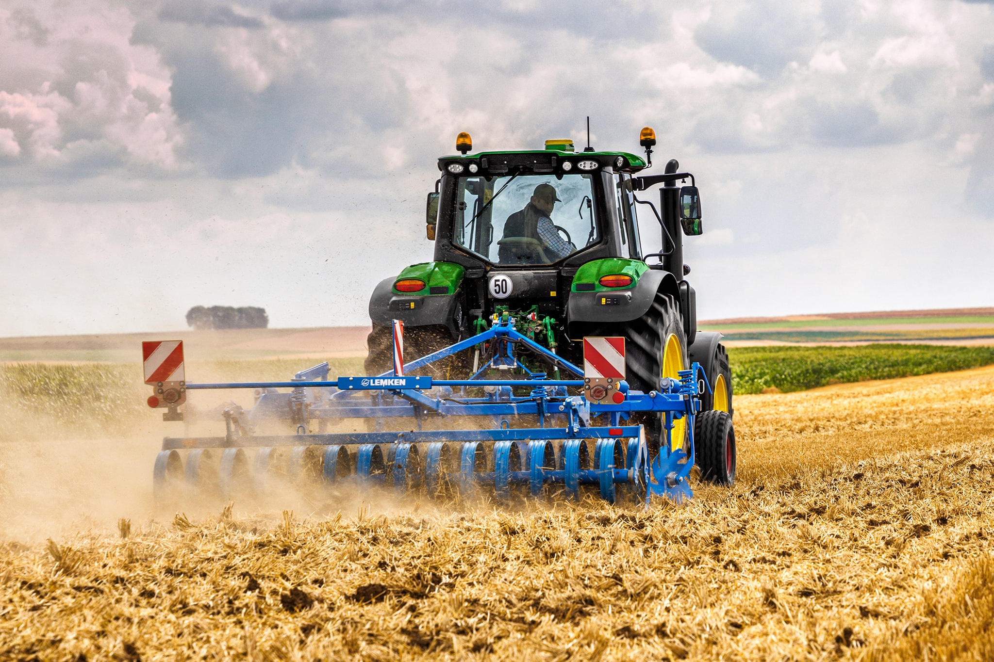 John Deere-traktor i 6M-serien som dyrker en innhøstet åker med en skålharv, sett bakfra under støvfulle åkerforhold.