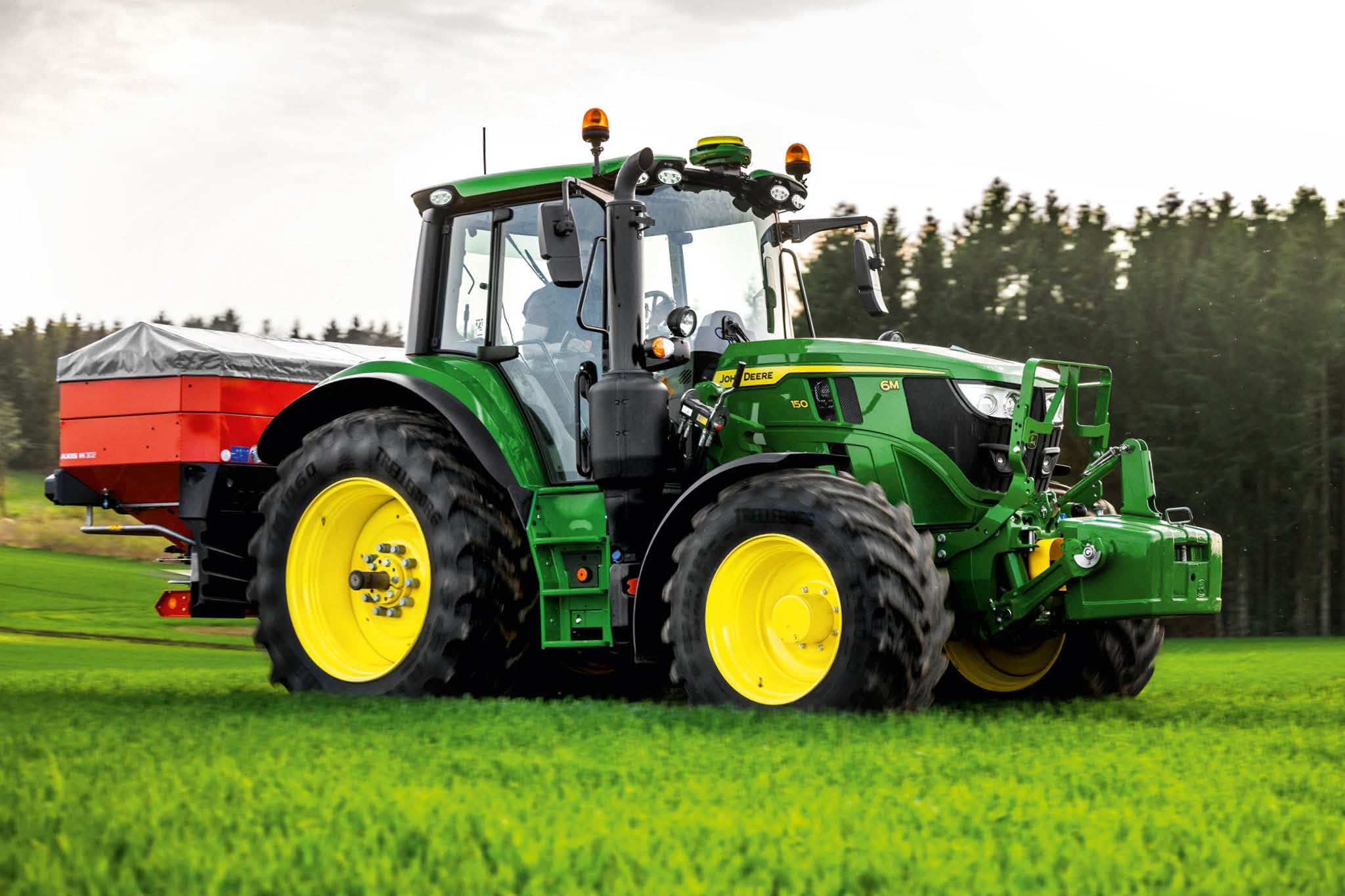 John Deere-traktor i 6M-serien med bakmontert sprederbom som arbeider på gress nær en skogskant.