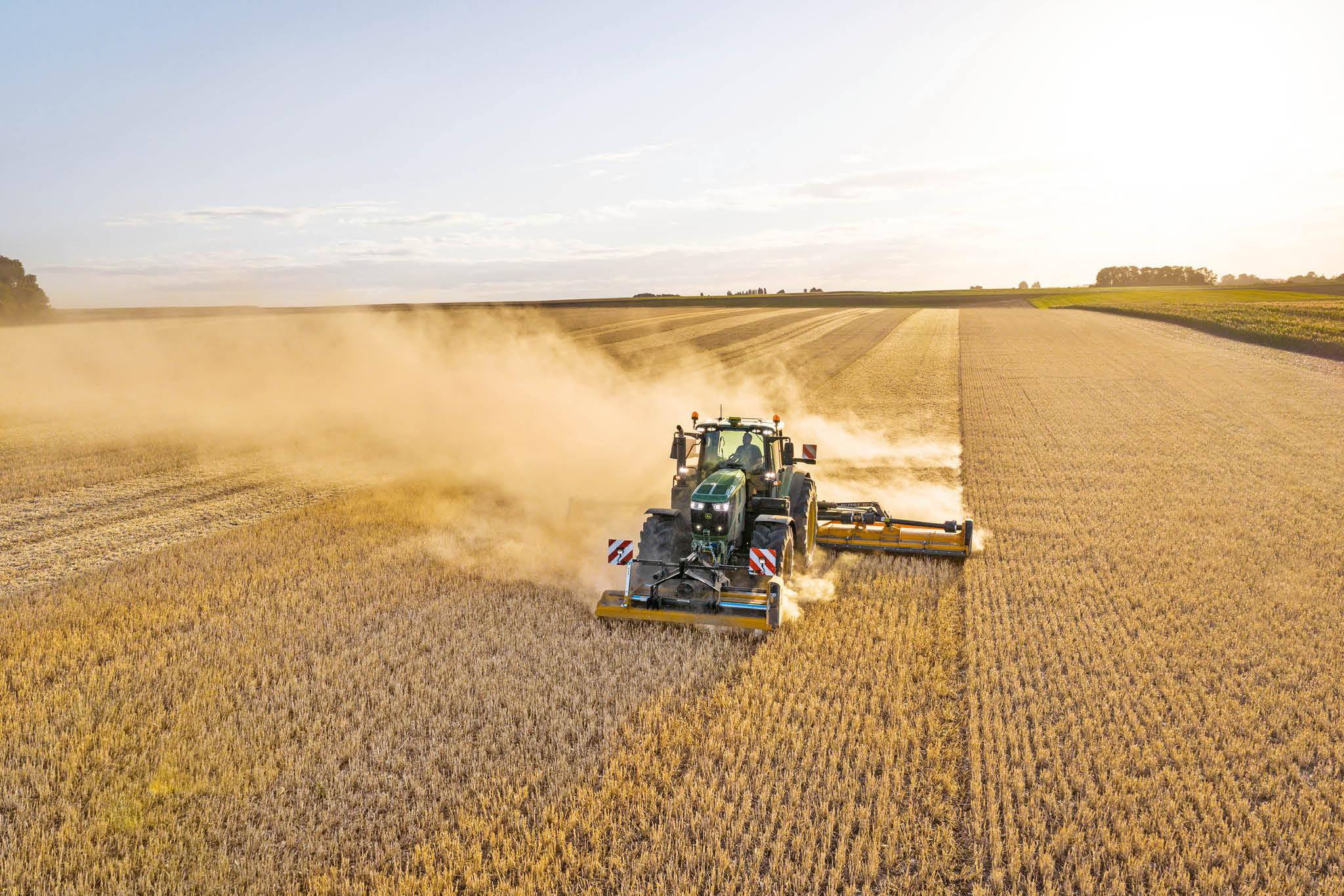 John Deere-traktor i 6M-serien tauer en gjødselvogn langs en landevei avgrenset av gress og åkre.