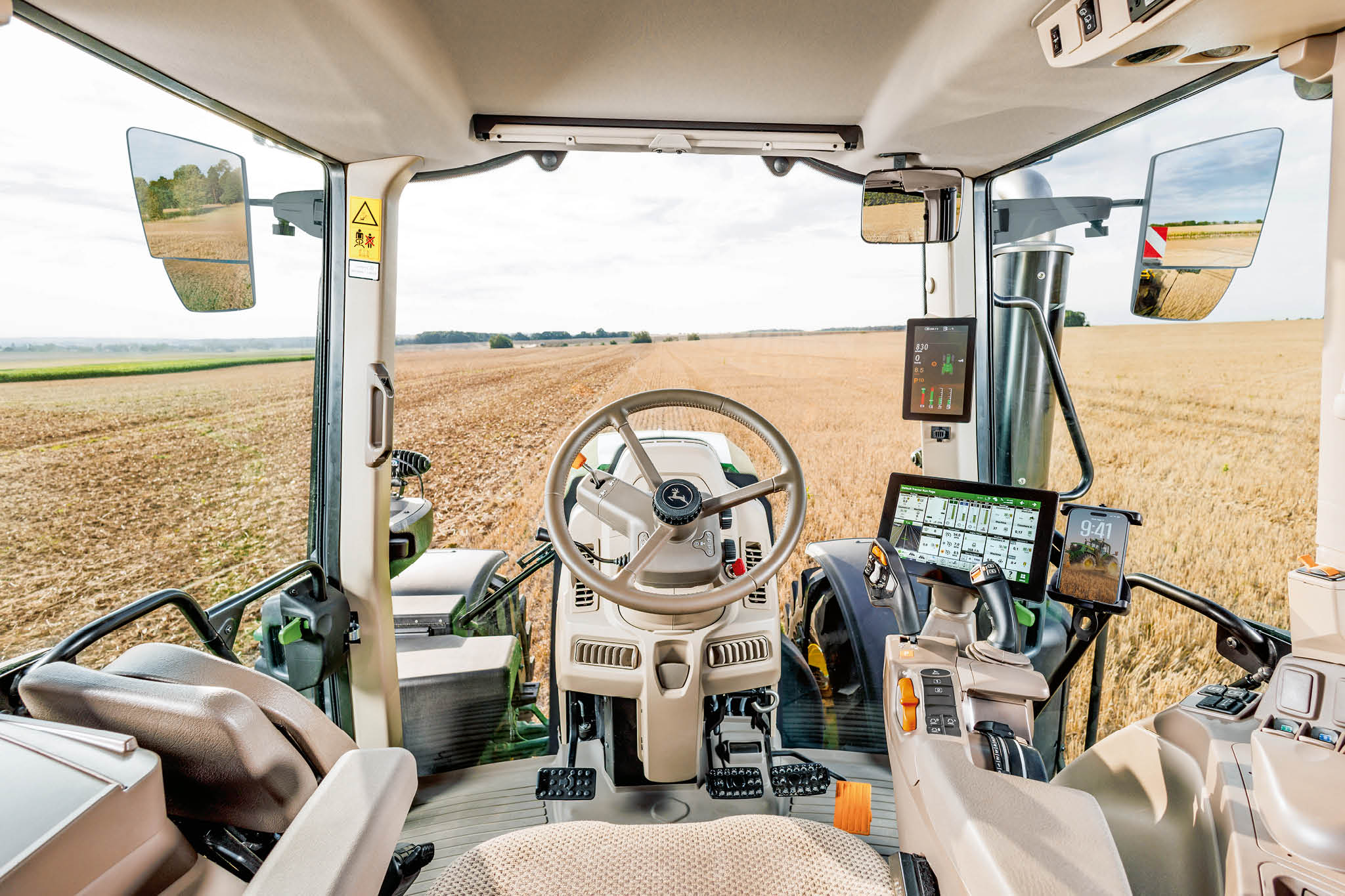 Sett fra innsiden av en John Deere-førerhytte i 6M-serien som viser ratt, kontrolldisplayer og stubbfelt foran.