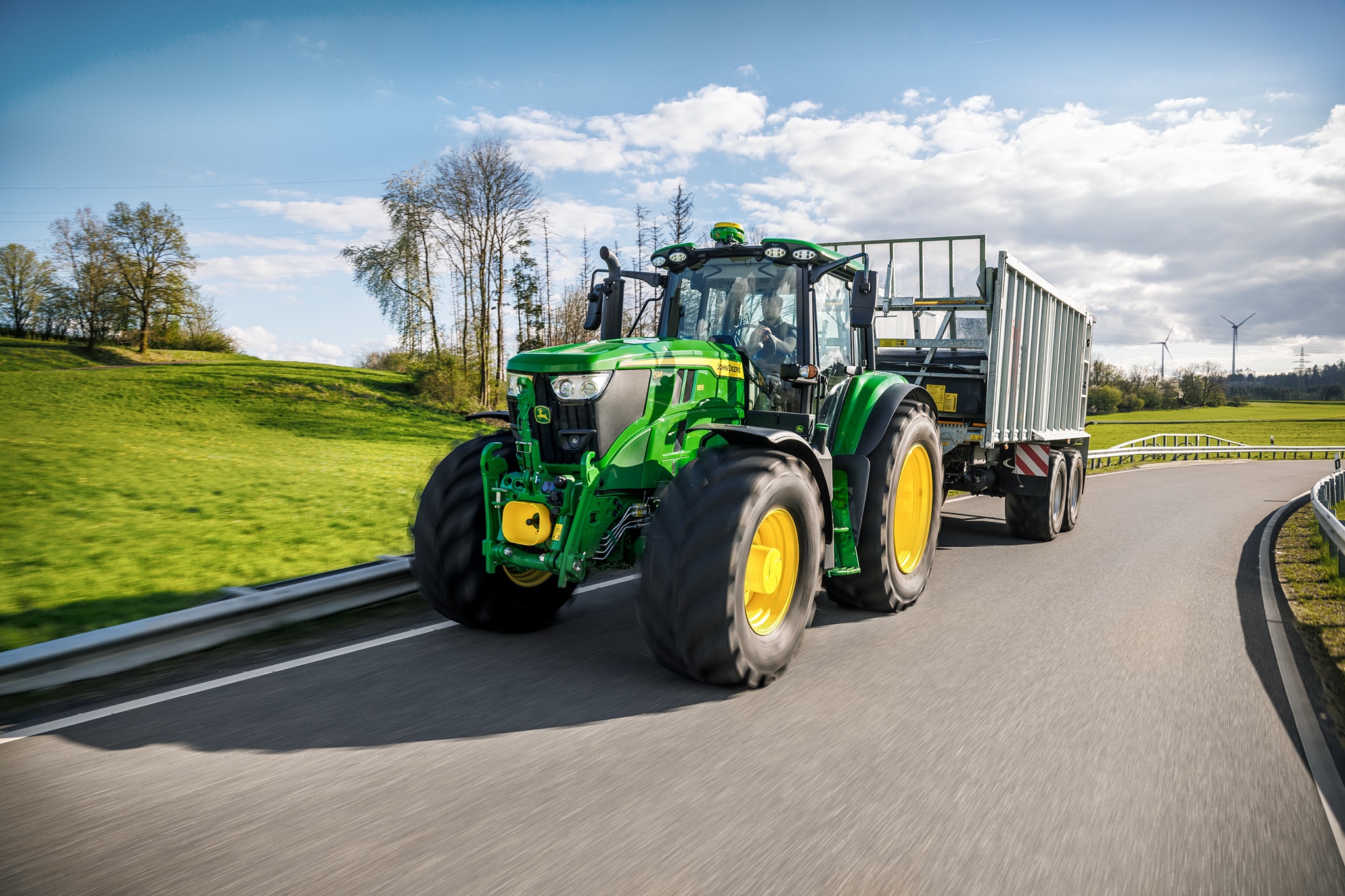John Deere-traktor i 6M-serien som transporterer en høysidig tilhenger på asfaltert vei.