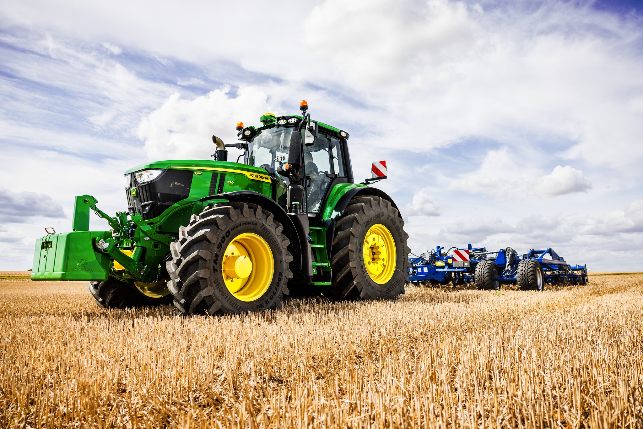 John Deere-traktor i 6M-serien som trekker et tungt pløyeredskap over en stubbåker under skydekke.