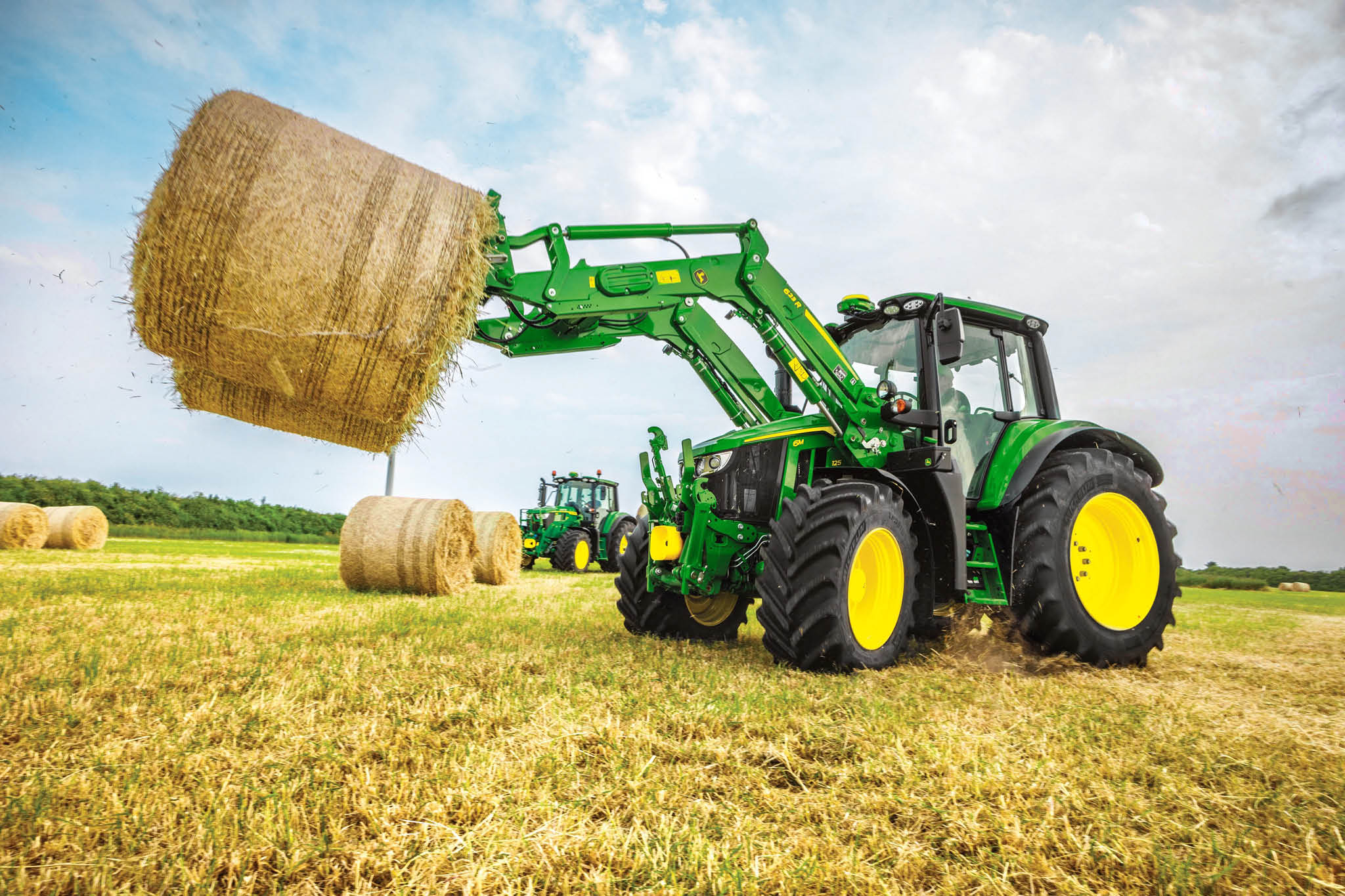 John Deere-traktor i 6M-serien løfter en stor rundballe med frontlaster på en åpen åker.