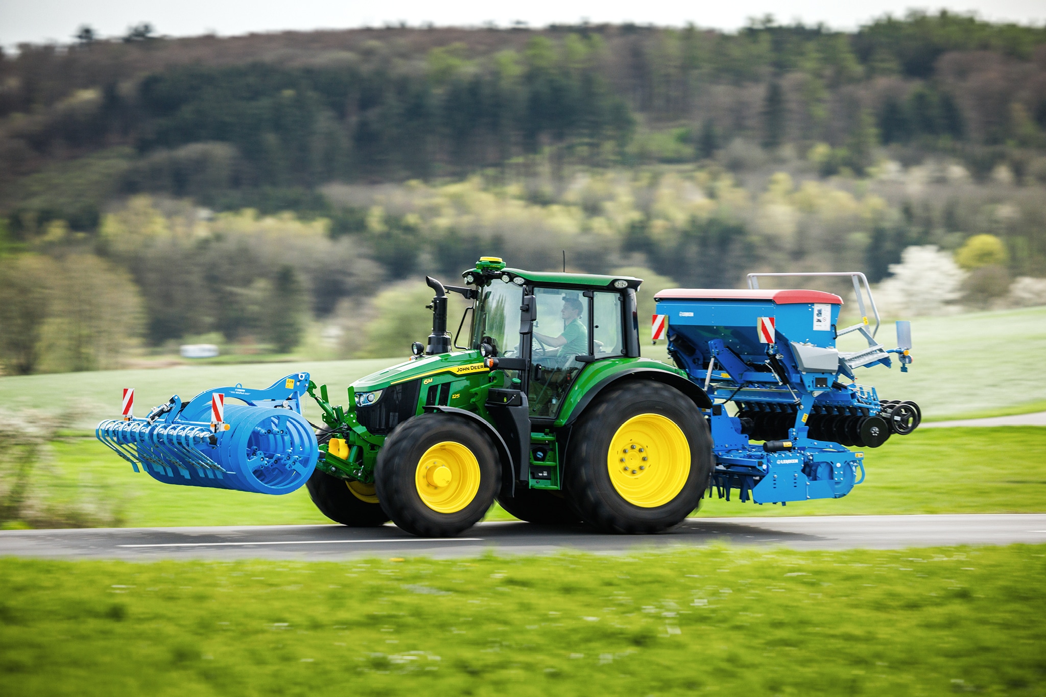 John Deere-traktor i 6M-serien som transporterer frontmontert redskap og bakre såmaskin langs en landevei.