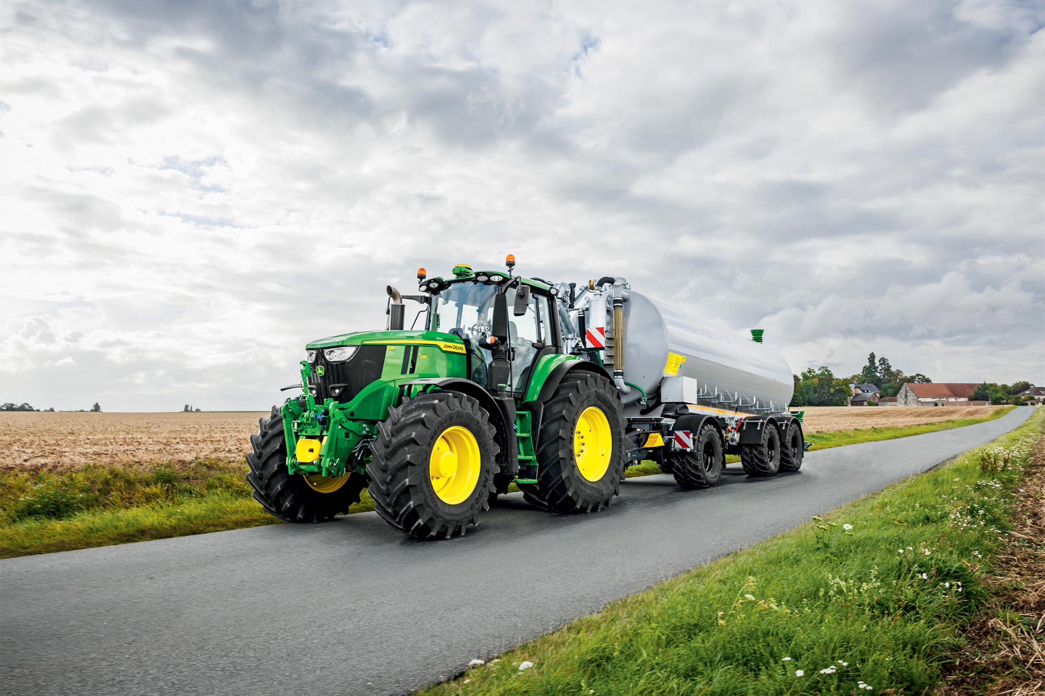 John Deere-traktor i 6M-serien som arbeider på en innhøstet åker med brede kultivatorer, sett ovenfra med støv som stiger opp.