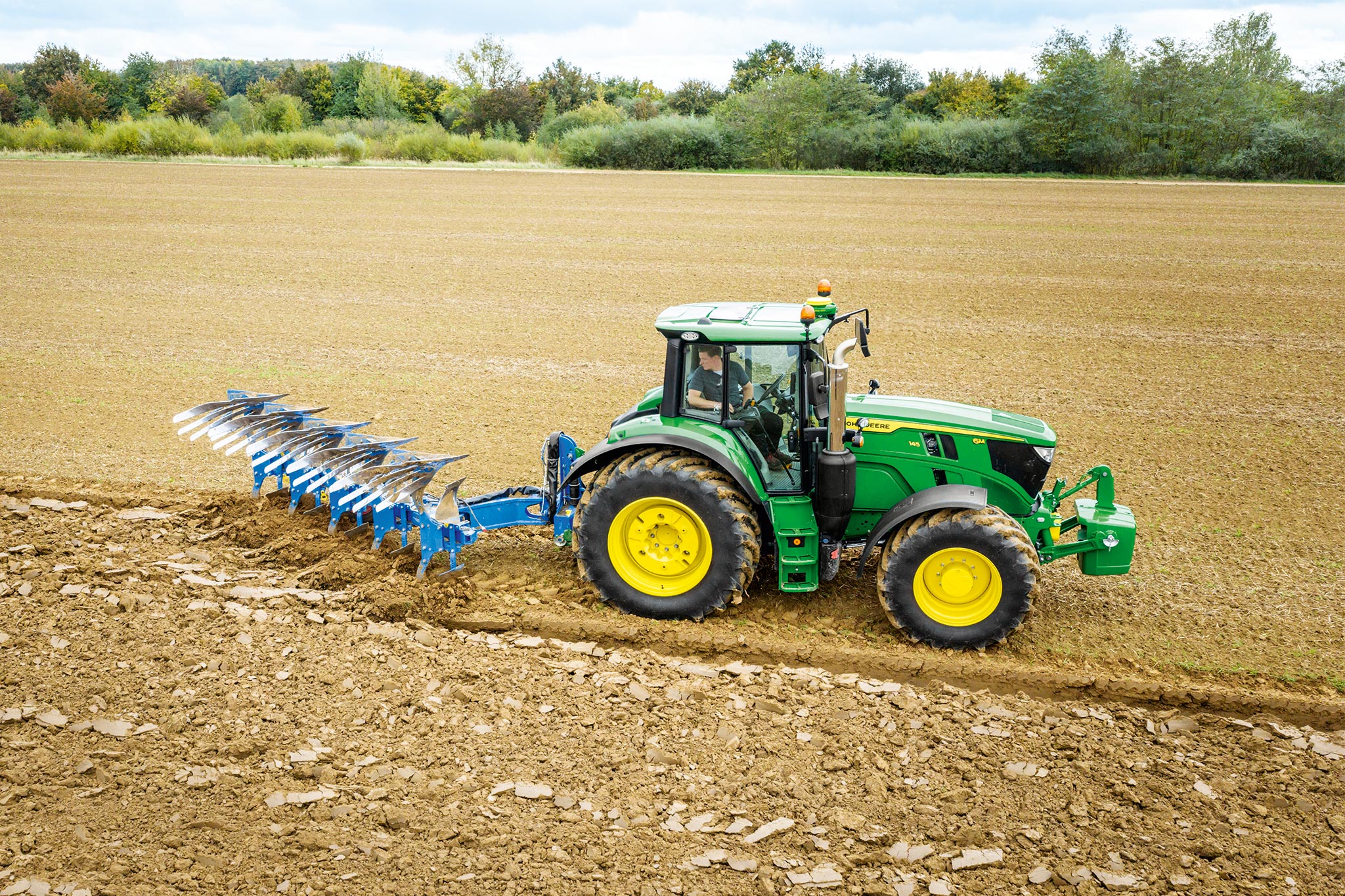 John Deere-traktor i 6M-serien som arbeider i et pløyd felt med et bakmontert pløyeredskap under overskyet himmel.