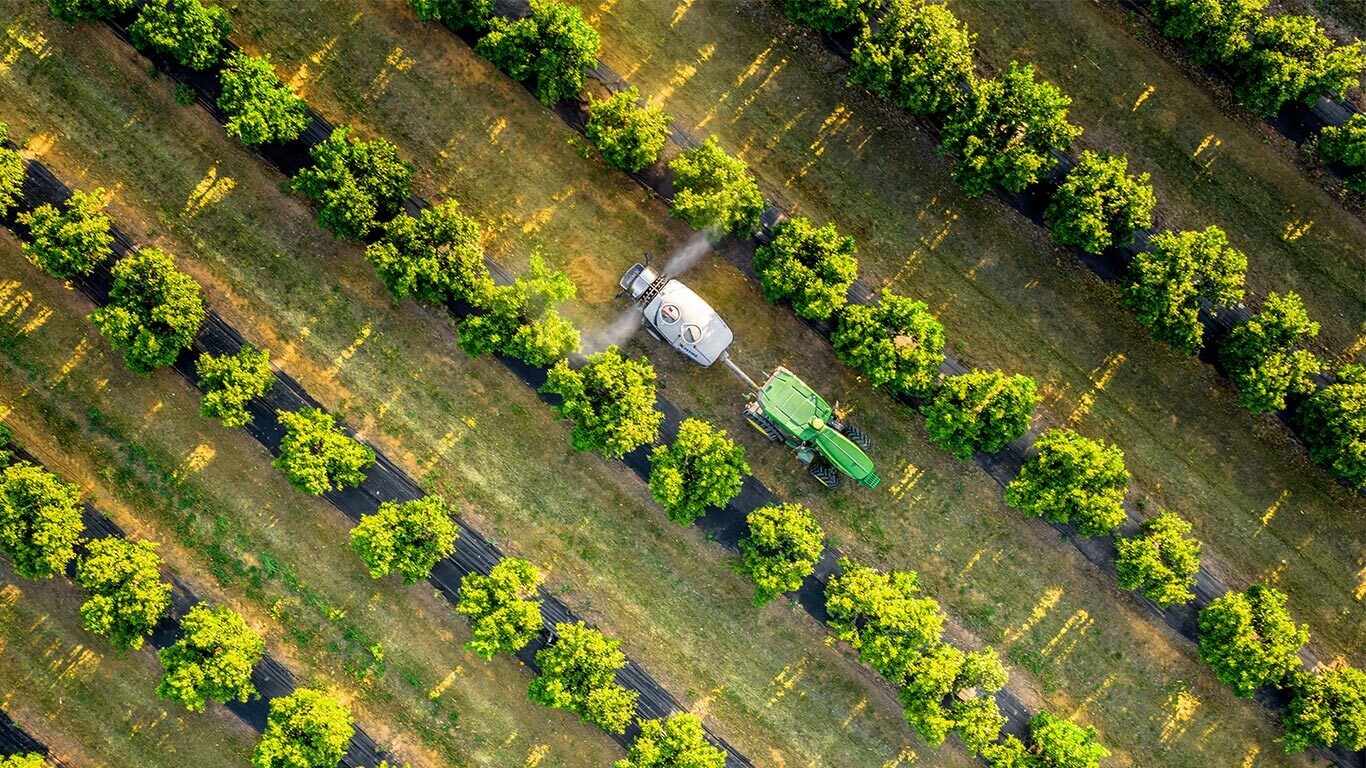 Flyfoto av en grønn og gul John Deere 5015EN-traktor som sleper en sprøyte gjennom frukthager-rader med jevn avstand.