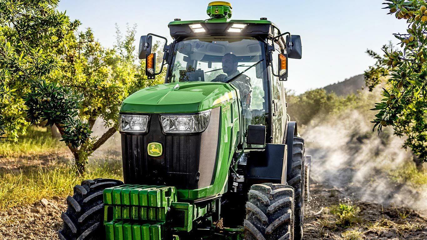 Grønn og gul John Deere 5075EN-traktor som kjører gjennom en frukthage på en jordbane, omgitt av frukttrær og sollys.