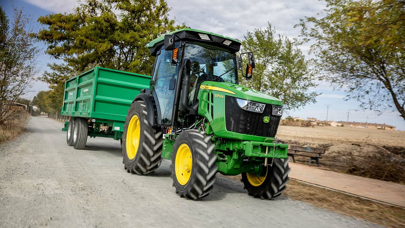 John Deere 5075EN traktor trekker en grønn tilhenger på en landlig grusvei med trær i bakgrunnen.