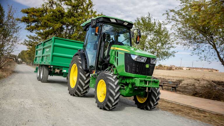 John Deere 5075EN traktor trekker en grønn tilhenger på en landlig grusvei med trær i bakgrunnen.