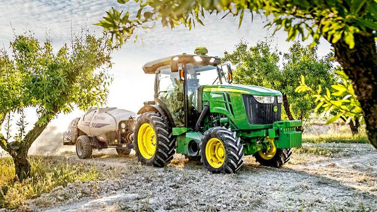 Grønn og gul John Deere 5090EN-traktor trekker en sprøyte gjennom en frukthage på en solrik dag.