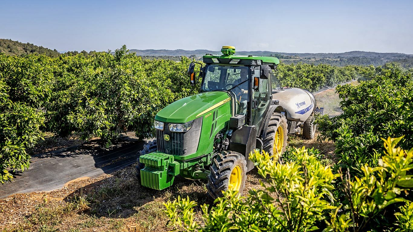 Grønn og gul John Deere 5105EN-traktor som tauer en sprøyte i en frodig frukthage med bølgende bakker i bakgrunnen.
