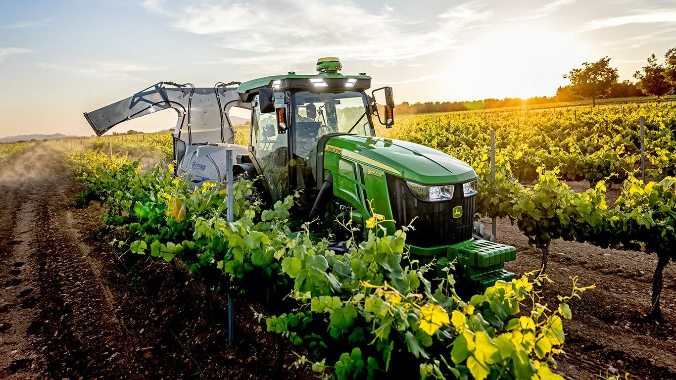 Grønn og gul John Deere 5105EN-traktor som arbeider i en vingård ved solnedgang, og sprøyter avlinger mellom radene av vinranker.