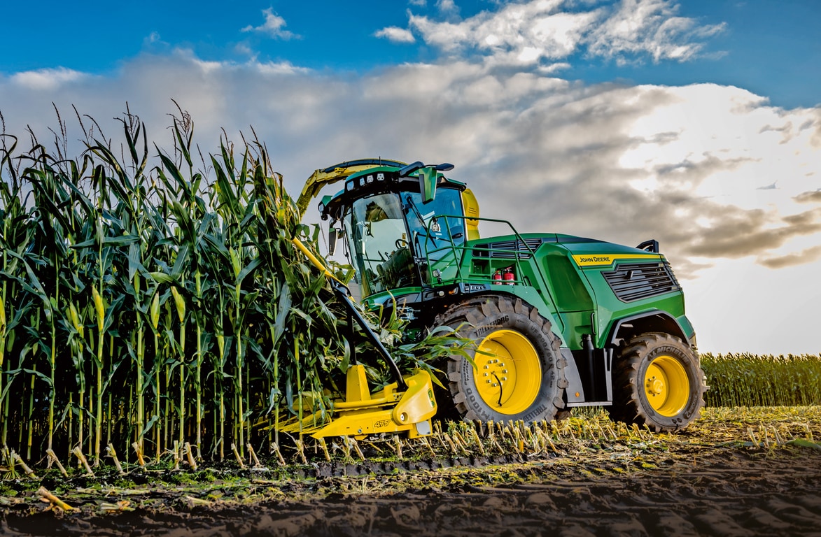 John Deere fôrhøster høster mais på en åker under blå himmel med skyer.