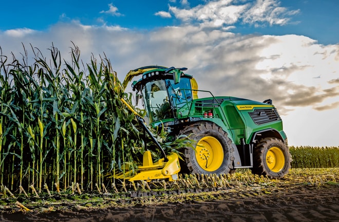 John Deere fôrhøster høster mais på en åker under blå himmel med skyer.