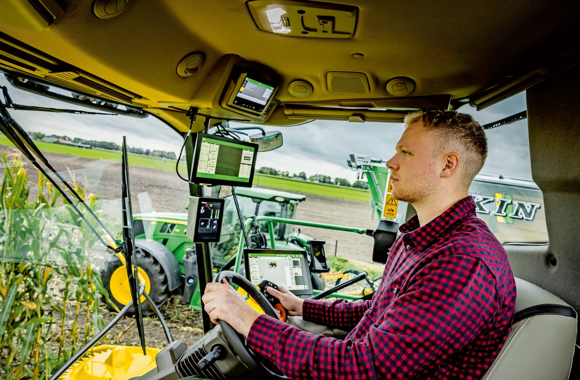 Bonde kjører avansert landbruksmaskineri med flere digitale skjermer inne i en selvgående fôrhøster, for å vise frem moderne landbruksteknologi på et felt.