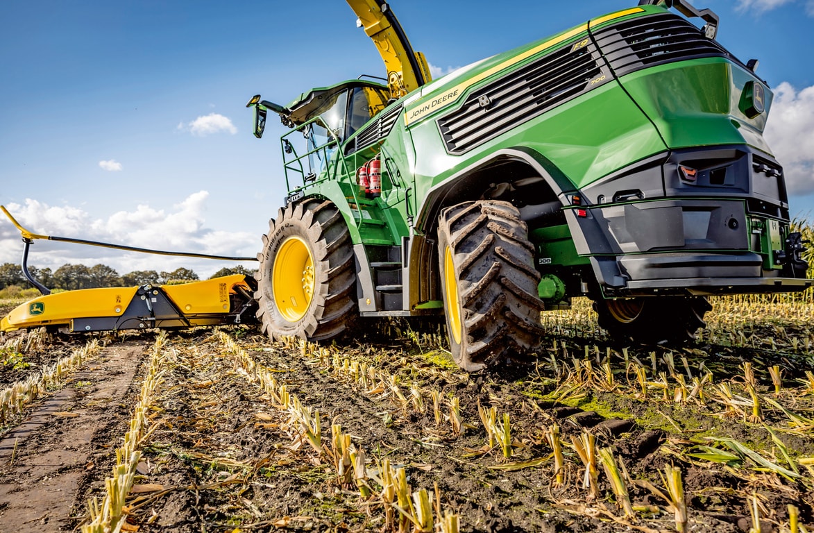 John Deere fôrhøster arbeider på en innhøstet maisåker, og viser maskinens store dekk og kuttemekanisme mot en klar blå himmel.