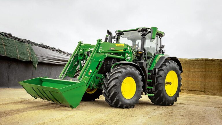 John Deere-traktor med frontlaster parkert på e gårdstun nær silofôrlagring.