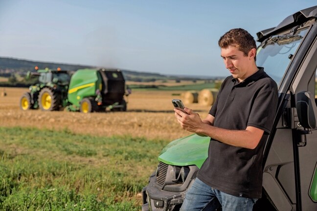 Personen som sjekker balledokumentasjonsdataene på mobiltelefonen med en John Deere VR-ballepresse i bakgrunnen